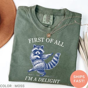May include: A moss-colored t-shirt with the text "FIRST OF ALL I'M A DELIGHT" and a blue illustration of a raccoon. The shirt has a crew neck and short sleeves. A gold necklace is draped across the shirt.