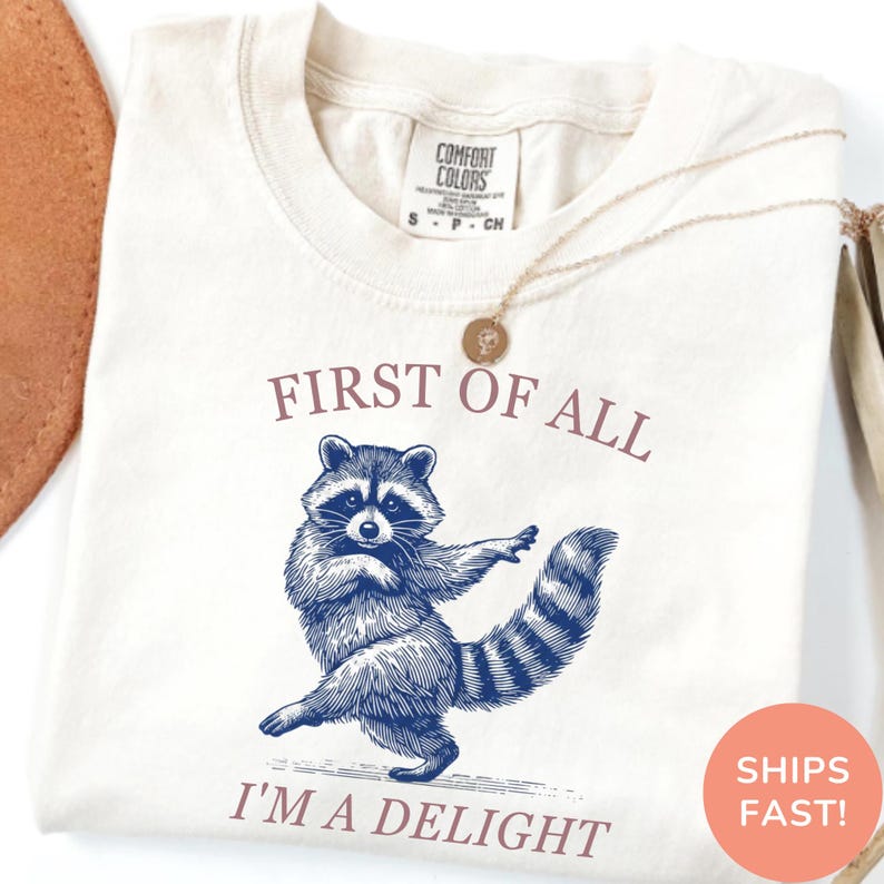 May include: Cream-colored t-shirt with a blue illustration of a raccoon and the text "FIRST OF ALL I'M A DELIGHT". The shirt has a crew neck and short sleeves. A gold necklace with a pendant is draped on the shirt.