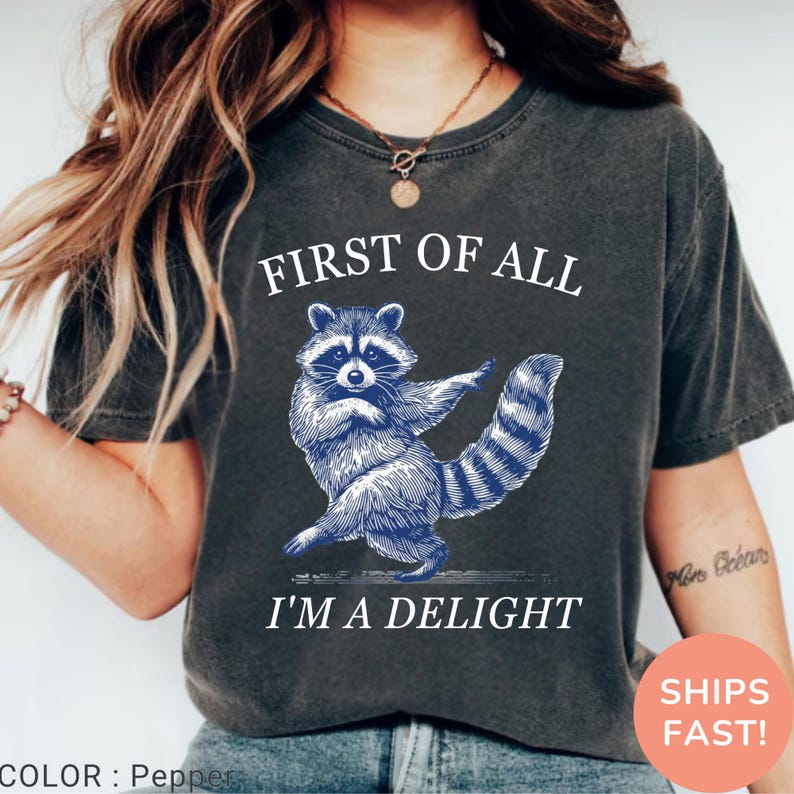 May include: Dark gray t-shirt featuring a blue illustration of a raccoon with the text "FIRST OF ALL I'M A DELIGHT". The raccoon is depicted in a dancing pose. The shirt is a solid color and has a crew neck.