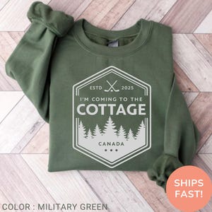 May include: Military green sweatshirt with a white graphic that reads "I'M COMING TO THE COTTAGE CANADA" inside a hexagon. The graphic includes crossed hockey sticks and a forest silhouette. The sweatshirt has long sleeves.