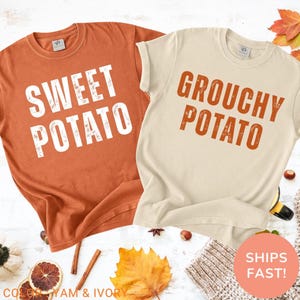 May include: Two t-shirts, one orange and one cream, with the words "Sweet Potato" and "Grouchy Potato" printed on them in large, distressed fonts. The shirts are displayed with fall-themed decorations, including leaves and cinnamon sticks.