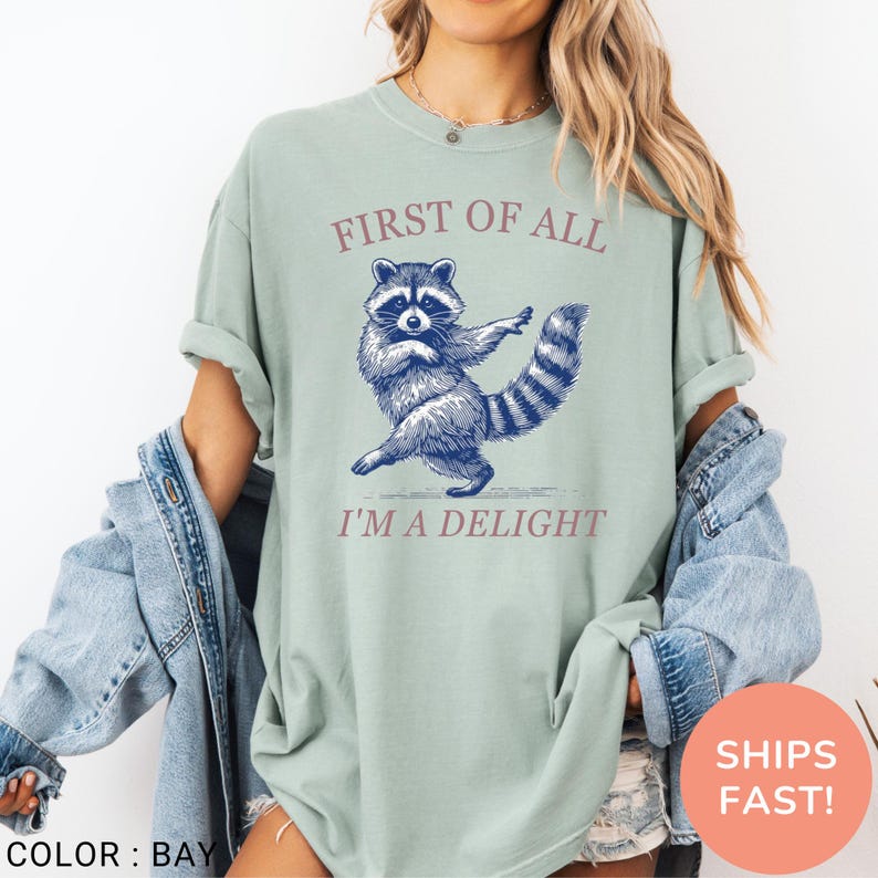May include: A light green t-shirt with a raccoon graphic and the text "FIRST OF ALL I'M A DELIGHT". The raccoon is depicted in blue, with a striped tail. The shirt is a relaxed fit.