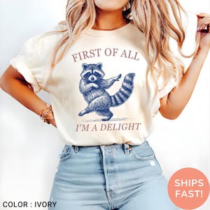 May include: Ivory-colored t-shirt featuring a blue raccoon illustration with the text "FIRST OF ALL I'M A DELIGHT". The raccoon is depicted in a dancing pose. The shirt is a casual wear item.