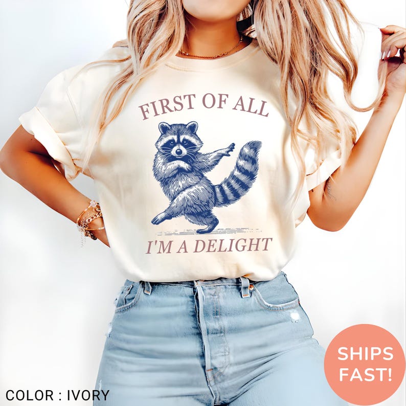 May include: Ivory-colored t-shirt featuring a blue raccoon illustration with the text "FIRST OF ALL I'M A DELIGHT". The raccoon is depicted in a dancing pose. The shirt is a casual wear item.