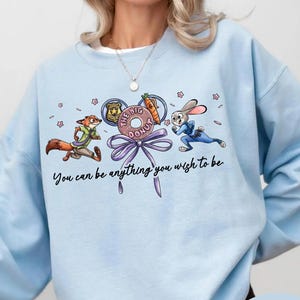 May include: Light blue sweatshirt featuring cartoon characters from Zootopia. The design includes Nick Wilde, Judy Hopps, and a donut with the text "The Big Donut". The phrase "You can be anything you wish to be" is also printed on the sweatshirt.