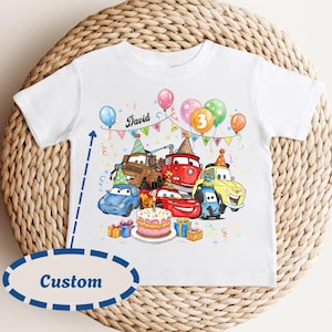 May include: White toddler t-shirt featuring a colorful birthday party design. The graphic includes cartoon cars, balloons, a cake, and the name "David". The number "3" is displayed on a balloon, suggesting a third birthday.
