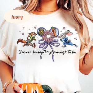 May include: Ivory-colored t-shirt featuring a cartoon design with a fox and a rabbit running, and a donut with the text "The Big Donut". The text "You can be anything you wish to be" is also printed on the shirt.