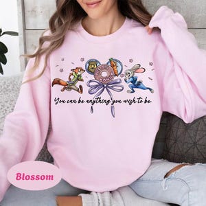 May include: Light pink sweatshirt with a cartoon graphic featuring a fox and a rabbit, with the text "You can be anything you wish to be." The graphic includes a donut and a carrot. The word "Blossom" is printed on the bottom.