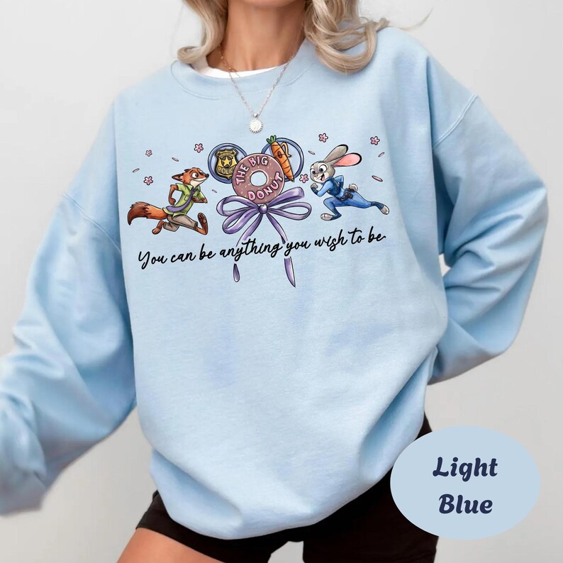 May include: Light blue sweatshirt featuring cartoon characters from Zootopia. The design includes Nick Wilde, Judy Hopps, and a donut with the text "The Big Donut". The phrase "You can be anything you wish to be" is also printed on the sweatshirt.