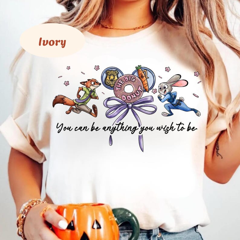 May include: Ivory-colored t-shirt featuring a cartoon design with a fox and a rabbit running, and a donut with the text "The Big Donut". The text "You can be anything you wish to be" is also printed on the shirt.