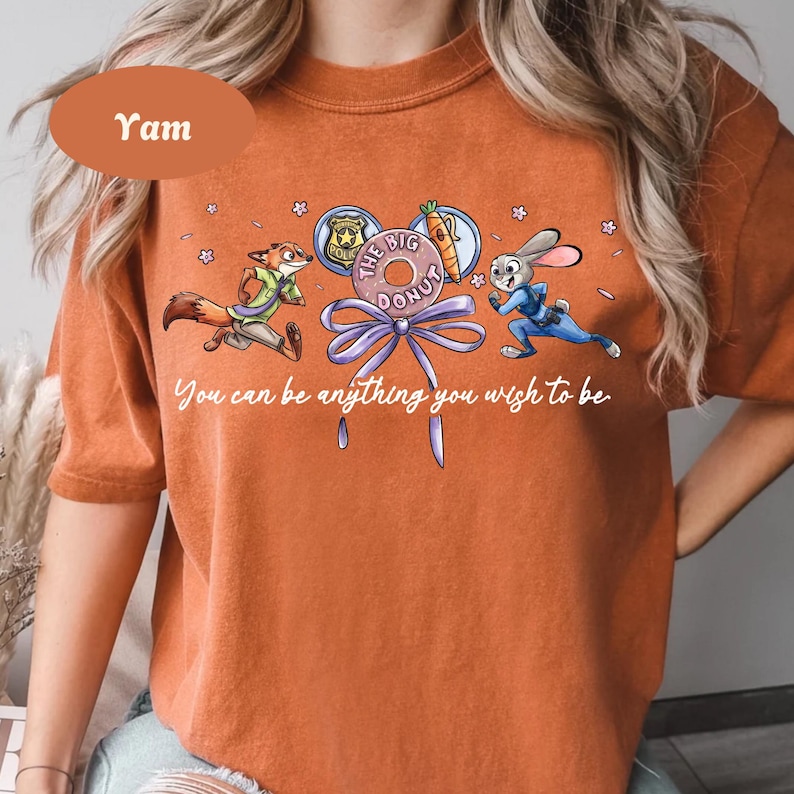 May include: Orange t-shirt featuring cartoon characters from Zootopia. The design includes Nick Wilde, Judy Hopps, and a donut with the text "The Big Donut." The shirt also has the text "You can be anything you wish to be."