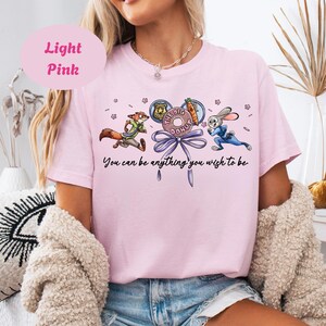 May include: Light pink t-shirt featuring cartoon characters from Zootopia. The design includes Nick Wilde, Judy Hopps, and a donut with the text "The Big Donut". The shirt also has the text "You can be anything you wish to be".