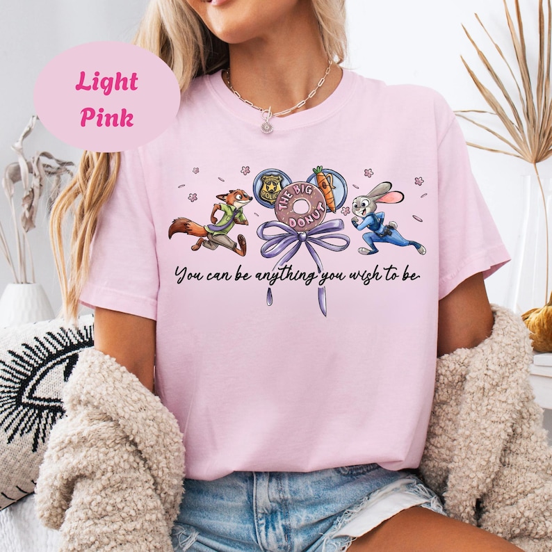 May include: Light pink t-shirt featuring cartoon characters from Zootopia. The design includes Nick Wilde, Judy Hopps, and a donut with the text "The Big Donut". The shirt also has the text "You can be anything you wish to be".