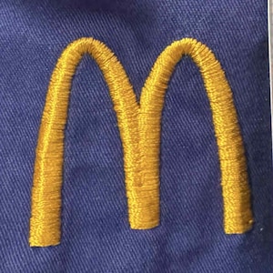 Puede incluir: Logotipo dorado bordado de McDonald's sobre tela azul. Los icónicos arcos dorados se muestran de forma destacada, mostrando el símbolo reconocible de la marca. La tela tiene una apariencia texturizada.