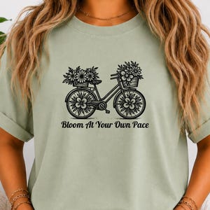 May include: A sage green short-sleeve t-shirt featuring a black graphic of a bicycle with flower baskets on the handlebars and rear rack, and flowers in the wheels. The text below the bicycle reads, Bloom At Your Own Pace.