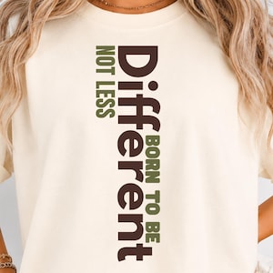 Op de afbeelding: Een crèmekleurig T-shirt met korte mouwen en verticale tekst in donkerbruin en olijfgroen. De tekst luidt "Different" in grote donkerbruine letters, met "BORN TO BE" en "NOT LESS" in een kleiner olijfgroen lettertype.