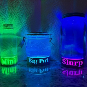 May include: Three illuminated glass containers with the words "Mini", "Big Pot", and "Slurp" glowing on their bases. The containers are lit with green, blue, and purple lights, creating a vibrant and colorful display. The jars are on a reflective surface.