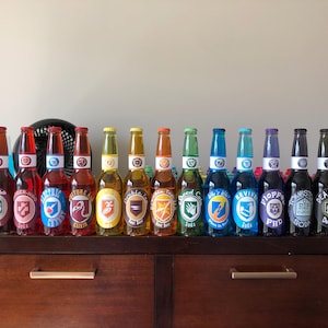 May include: A display of glass bottles with vibrant labels and caps, presented on a wooden cabinet. Each bottle showcases a distinct design and text, with a range of colours such as red, blue, yellow, and green. The bottles are of a consistent size and shape.