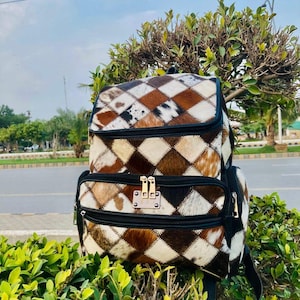 Peut inclure: Sac à dos en cuir de vachette multicolore avec un motif en losanges. Il est orné d'un rabat supérieur, d'une poche avant zippée et de bretelles noires. Les couleurs sont le marron, le blanc et le noir.