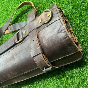 May include: A dark brown leather knife roll with a shoulder strap. The roll is secured with straps and brass-colored hardware. The leather has a distressed appearance, and the roll is partially open, revealing the interior.