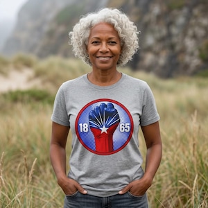 Op de afbeelding: Een grijs gemêleerd T-shirt met korte mouwen en een cirkelvormige afbeelding op de borst. Het ontwerp toont een opgeheven vuist in rood, wit en blauw met een witte ster, geflankeerd door de cijfers 18 en 65 in een cirkel met rode rand.