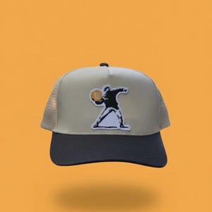 May include: A beige and black trucker hat with a Bitcoin-themed graphic. The hat features a black peak, a beige front panel with a patch of a person throwing a Bitcoin, and a mesh back.