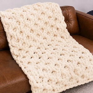May include: A chunky knit throw blanket in a solid cream color. The blanket features a thick, hand-knitted texture with large, interconnected loops throughout the entire surface, creating a soft and voluminous appearance across the fabric.