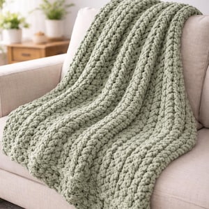 Puede incluir: Una manta de punto grueso verde salvia sobre un sofá beige. La manta tiene un patrón texturizado y acanalado y parece suave y acogedora. El sofá está en un salón bien iluminado.