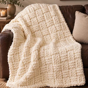 Peut inclure: Un jeté de lit en tricot épais de couleur crème blanc cassé présentant un motif de vannerie proéminent. La couverture est fabriquée à partir de boucles de fil épaisses et douces qui créent un aspect texturé en forme de grille.