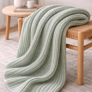 May include: A pale green knitted throw blanket draped over a wooden chair. The blanket has a ribbed texture and is made of a soft material. The chair is made of woven wicker and wood. The scene is set in a cosy living room.