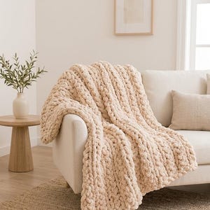 Puede incluir: Una manta de punto grueso en color crema sólido, con una construcción de hilo grueso y texturizado con grandes bucles visibles. La manta está colocada sobre el brazo y el asiento de un sofá de color claro, mostrando su apariencia suave y mullida.