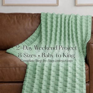 May include: A light green knitted blanket draped over a brown leather sofa. The blanket has a textured, woven pattern. Text on the blanket reads "2-Day Weekend Project, 8 Sizes, Baby to King, Complete Step-by-Step instructions."