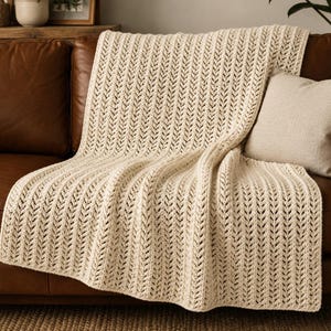 May include: A cream-colored crochet throw blanket featuring a repeating vertical herringbone stitch pattern. The blanket is draped over a brown leather sofa and is accompanied by a single square throw pillow in a matching cream-colored fabric.