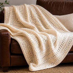 May include: A cream-coloured, crocheted blanket draped over a brown leather armchair. The blanket has a textured, open weave design. A matching cushion rests on the chair. The scene suggests a cosy, comfortable living space.