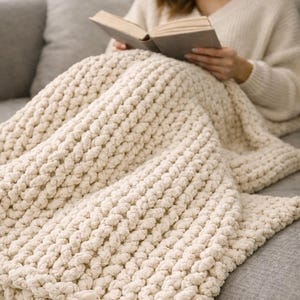May include: A cream-coloured, chunky knit blanket draped over a grey sofa. The blanket's thick texture is visible, with large, looped stitches creating a cosy appearance. A person is reading a book under the blanket.