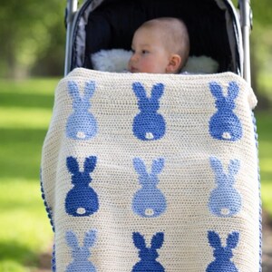 May include: A baby in a stroller is covered with a cream-colored crocheted blanket. The blanket features a pattern of blue bunny silhouettes. The stroller is black with a black canopy and is on a path with green grass in the background.