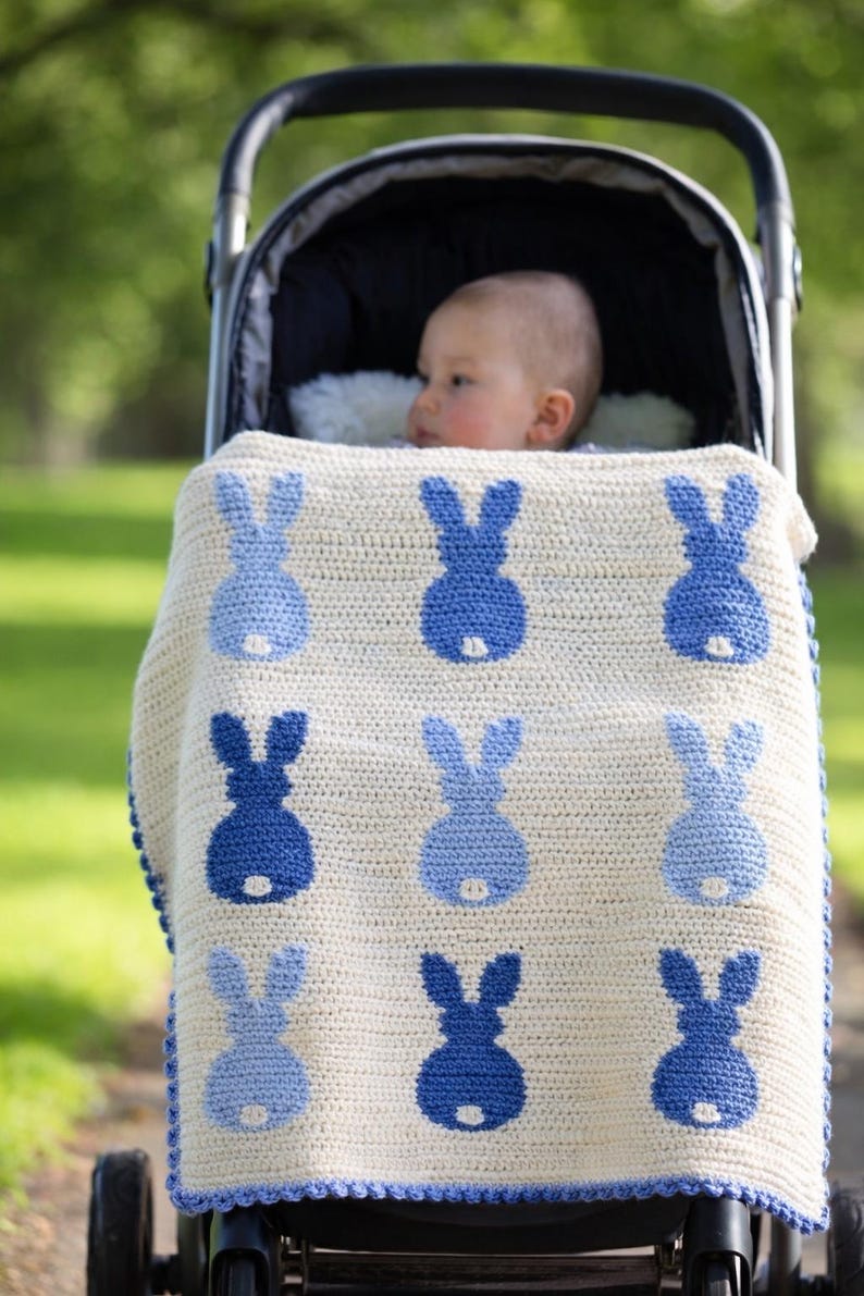May include: A baby in a stroller is covered with a cream-colored crocheted blanket. The blanket features a pattern of blue bunny silhouettes. The stroller is black with a black canopy and is on a path with green grass in the background.