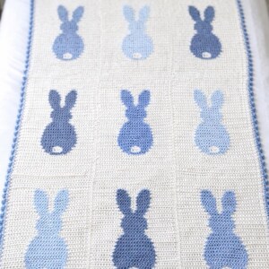 May include: A white and blue crochet blanket featuring a pattern of nine blue bunny silhouettes. The blanket has a blue scalloped border and is displayed on a bed. The bunny shapes vary in shades of blue.