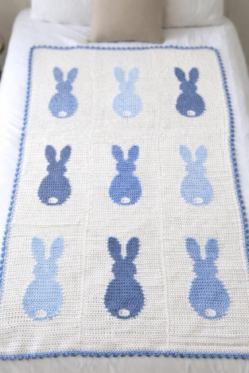 May include: A white and blue crochet blanket featuring a pattern of nine blue bunny silhouettes. The blanket has a blue scalloped border and is displayed on a bed. The bunny shapes vary in shades of blue.