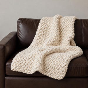 Peut inclure: Un plaid en tricot épais de couleur crème blanc cassé. La couverture présente un motif de tissage tressé et épais créé avec de grandes boucles de fil, offrant un aspect texturé sur toute la surface de cet accessoire textile.