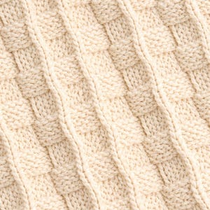 May include: Close-up of a cream-colored knitted fabric with a basketweave pattern. The texture is visible, showing the individual stitches and the diagonal lines created by the weave. The fabric appears soft and warm.