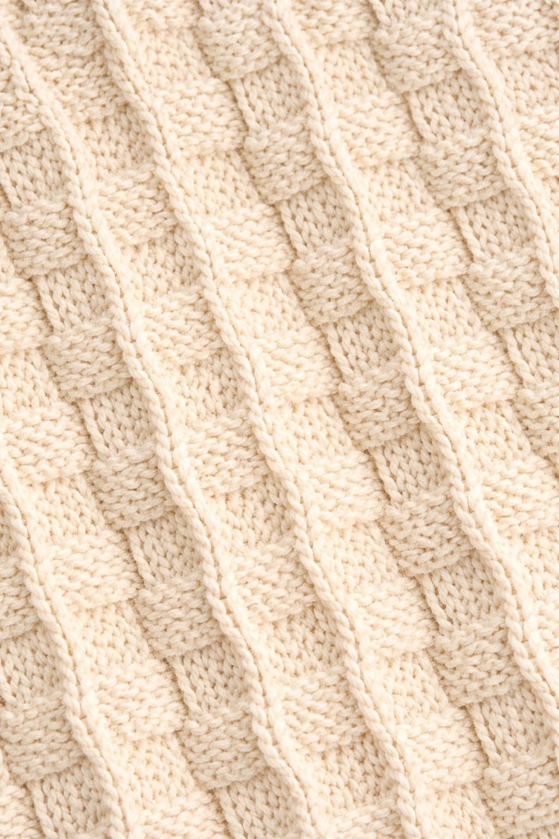 May include: Close-up of a cream-colored knitted fabric with a basketweave pattern. The texture is visible, showing the individual stitches and the diagonal lines created by the weave. The fabric appears soft and warm.