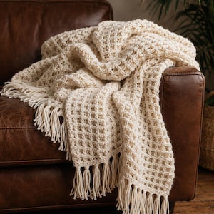 Peut inclure: Un jeté de lit couleur crème présentant un motif en maille gaufrée texturée et des détails à longues franges sur les bords. Le tissu a une apparence épaisse et tissée avec une structure en forme de grille sur toute la surface du textile.