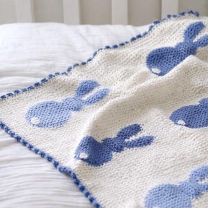 May include: A white and blue knitted baby blanket featuring a repeating pattern of blue bunny silhouettes. The blanket has a scalloped blue border and is displayed on a white bed.