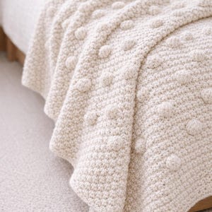 May include: A cream-coloured knitted blanket with a textured, raised dot pattern. The blanket is draped over a bed, showcasing its soft, cosy appearance. The neutral colour and tactile design suggest a focus on comfort and home decor.