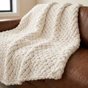 May include: A chunky knit throw blanket in an off-white cream color. The blanket features a thick, braided, or woven texture created with large, soft yarn loops. The material has a plush, voluminous appearance with a consistent pattern throughout.