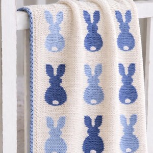 May include: A cream-colored, crocheted baby blanket featuring a pattern of blue bunny silhouettes. The blanket has a blue border and is draped over a white crib rail. The bunny shapes are in two shades of blue.