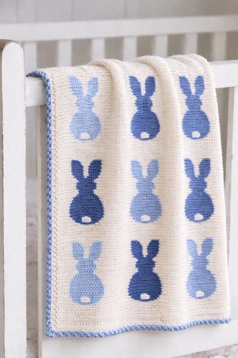 May include: A cream-colored, crocheted baby blanket featuring a pattern of blue bunny silhouettes. The blanket has a blue border and is draped over a white crib rail. The bunny shapes are in two shades of blue.
