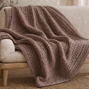 May include: A dusty rose crochet throw blanket featuring a textured pattern of alternating vertical ribbed columns and intricate openwork lace sections. The blanket is draped over a light-colored sofa, showcasing its soft yarn construction.