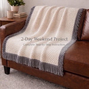 May include: A cream-colored crocheted blanket with a gray border and fringe is draped over a brown leather couch. The blanket has a textured pattern and the text "2-Day Weekend Project" is visible on the blanket.